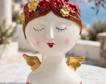 May include: A ceramic head-shaped planter featuring a white face with closed eyes, pink cheeks, and red lips. The figure has brown hair adorned with red and gold flowers, and small gold wings on the shoulders. The base is painted in a solid red color.