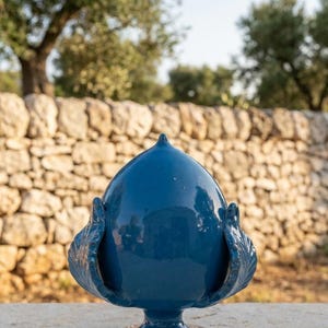 Può includere: Una scultura in ceramica a forma di pigna di colore blu intenso con una finitura lucida e riflettente. L'oggetto presenta una sommità appuntita, una base a piedistallo e decorazioni laterali stilizzate a forma di foglia su entrambi i lati.