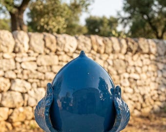 May include: A glossy, deep blue ceramic pinecone sculpture featuring a pointed top and a pedestal base. The object is adorned with stylized, leaf-like scales on both sides, showcasing a smooth, reflective finish and a solid, uniform color throughout.