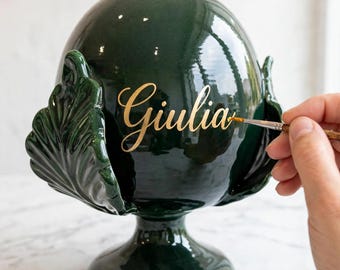 May include: A dark green ceramic decorative object shaped like a pinecone with a pedestal base and leaf-like accents. The name Giulia is hand-painted in gold script lettering on the front of the glossy surface, with a fine brush applying the paint.