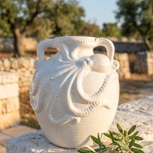 Peut inclure: Un vase en céramique blanc brillant orné d'un motif sculptural en relief représentant une pieuvre enroulée autour de l'extérieur. Le récipient possède deux anses symétriques intégrées aux tentacules, une large ouverture et un corps arrondi.