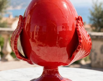 May include: A glossy, bright red ceramic decorative object shaped like a pinecone or egg, featuring stylized leaf-like protrusions on the sides. The piece stands on a circular pedestal base with a smooth, reflective finish and a pointed top.