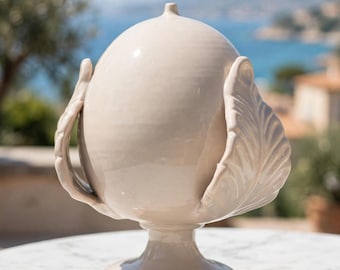 May include: A glossy, light beige ceramic decorative urn featuring a smooth, rounded central body topped with a small finial. The piece is supported by a pedestal base and is flanked by two symmetrical, sculpted leaf-shaped handles.