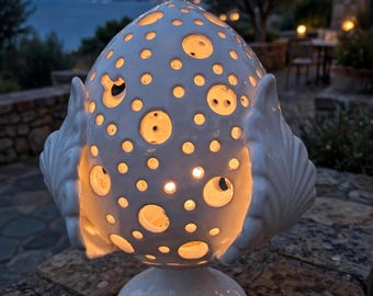 May include: A white ceramic lamp with a rounded, perforated body featuring numerous circular cutouts of varying sizes. The lamp is flanked by two sculpted, wing-like side elements and rests on a circular base, emitting a warm, soft interior glow.