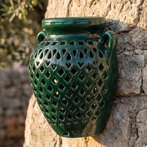 May include: A dark green glazed ceramic wall planter featuring a classic amphora shape with two side handles. The body of the vessel is decorated with a repeating lattice pattern of cut-out diamond shapes, allowing light to pass through the structure.