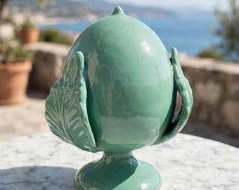 May include: A glossy, light green ceramic ornament shaped like a pinecone or artichoke, featuring a smooth, rounded central body and stylized, textured leaf-like protrusions at the base, all supported by a circular pedestal stand.