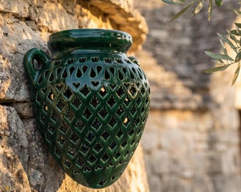 May include: A dark green ceramic wall planter with a glossy finish and a lattice cutout pattern. The vessel features a wide circular opening at the top, two small side handles, and a tapered base, designed to be mounted flush against a vertical surface.