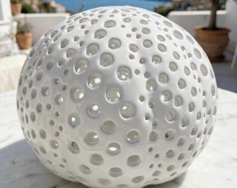 May include: A spherical white ceramic object featuring a dense, irregular pattern of circular cutouts of varying sizes across its entire surface. The glossy, smooth finish reflects light, creating a textured appearance reminiscent of a sea urchin shell.