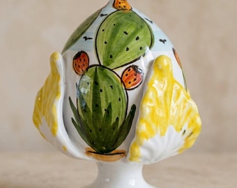 May include: A ceramic pinecone-shaped decorative object featuring hand-painted green cactus motifs with orange prickly pears on a light blue background. The base is a solid blue ring, and the sides are adorned with bright yellow, leaf-like protrusions.