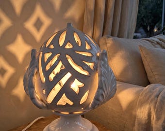 May include: A white ceramic table lamp shaped like a pinecone with intricate cut-out patterns that allow light to shine through. The lamp features a rounded, textured body with leaf-like details and a circular base with a white power cord.