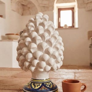 Può includere: Una scultura in ceramica bianca a forma di pigna con finitura lucida, dotata di una base decorata con motivi geometrici gialli, blu e neri, posizionata accanto a una piccola tazza in ceramica color terracotta.