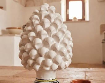 May include: A white ceramic pinecone sculpture with a glossy finish, featuring a base decorated with yellow, blue, and black geometric patterns, positioned next to a small, plain terracotta-colored ceramic cup on a textured stone surface.