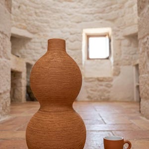 Può includere: Un vaso a forma di zucca color terracotta con una superficie testurizzata e striata, posizionato accanto a un set di tazzina da espresso e piattino in terracotta abbinati, con una finitura interna bianca e liscia.