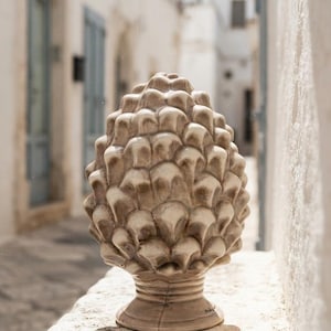 Puede incluir: Una escultura de cerámica con forma de piña que presenta un esmalte brillante de color crema con sombreado marrón en los huecos de las escamas. El objeto tiene una parte superior redondeada y texturizada sobre una base circular de pedestal.