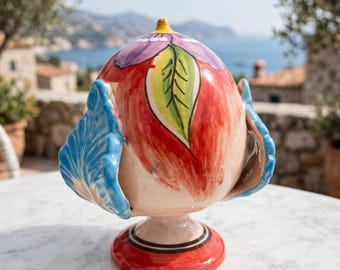 May include: A ceramic pigna ornament featuring a cream-colored base with hand-painted red, purple, and green floral motifs. The piece has two prominent blue leaf-shaped side handles and rests on a circular red pedestal base with black ring details.