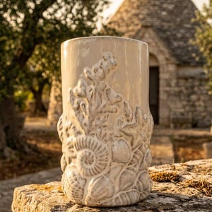 Könnte beinhalten: Eine zylindrische Keramikvase mit glänzend cremeweißer Oberfläche, die mit einem erhabenen, strukturierten Relief aus verschiedenen Muscheln, Korallen und Meereslebewesen verziert ist, das die Außenseite von der Basis bis zum oberen Rand bedeckt.