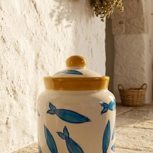Può includere: Un barattolo cilindrico in ceramica con base color crema, caratterizzato da motivi di pesci blu dipinti a mano sull'esterno. Il barattolo include un coperchio coordinato color crema sormontato da un pomello giallo e un bordo giallo alla base.