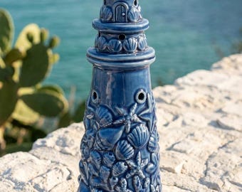 May include: A glossy blue ceramic lighthouse figurine featuring a textured surface decorated with relief carvings of various seashells, starfish, and coral. The structure is topped with a small starfish and includes small circular openings.