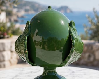 May include: A glossy, dark green ceramic decorative object shaped like a pinecone, featuring a rounded top, a pedestal base, and stylized leaf-like protrusions extending from the sides, creating a symmetrical and smooth, reflective surface.