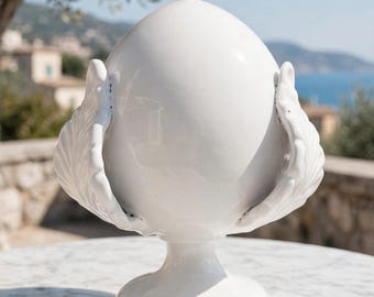 May include: A glossy white ceramic decorative object shaped like a large egg or pinecone, featuring sculpted leaf-like protrusions on both sides. The piece stands on a circular pedestal base and has a smooth, reflective finish throughout.