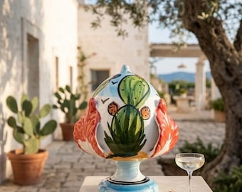 Puede incluir: Un adorno de cerámica pigna con un diseño de cactus verde pintado a mano sobre un fondo azul claro, acentuado con vibrantes protuberancias rojas en forma de hoja en los lados y una base circular, sobre un pedestal de piedra junto a una copa.