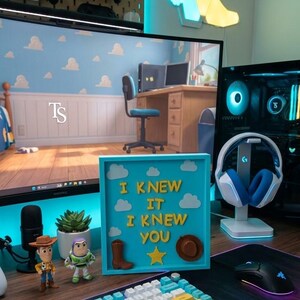 Pu&ograve; includere: Una cornice quadrata azzurra tipo shadow box con il testo giallo I KNEW IT I KNEW YOU, ritagli di nuvole bianche, uno stivale da cowboy marrone, un cappello da cowboy marrone e una stella gialla, posizionata su una scrivania accanto a due statuine.