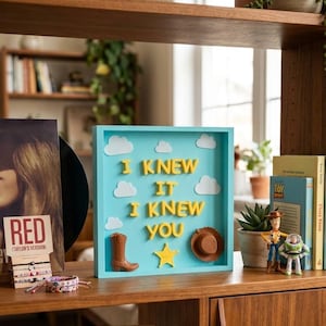 Pu&ograve; includere: Una scatola quadrata azzurra con il testo in rilievo giallo I KNEW IT I KNEW YOU. L'espositore include ritagli di nuvole bianche, uno stivale da cowboy marrone, una stella gialla e un cappello da cowboy marrone fissati sullo sfondo azzurro.