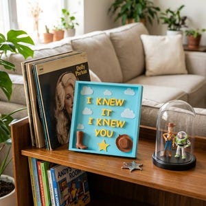 Pu&ograve; includere: Una cornice quadrata azzurra contenente lettere gialle in 3D che formano la scritta I KNEW IT I KNEW YOU, decorata con nuvole in feltro bianco, uno stivale da cowboy marrone, un cappello da cowboy marrone e una stella gialla.