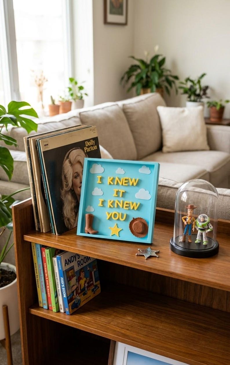 Pu&ograve; includere: Una cornice quadrata azzurra contenente lettere gialle in 3D che formano la scritta I KNEW IT I KNEW YOU, decorata con nuvole in feltro bianco, uno stivale da cowboy marrone, un cappello da cowboy marrone e una stella gialla.