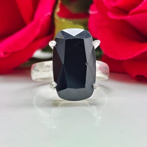 Könnte beinhalten: Ein Silberring mit einem großen, rechteckigen, facettierten schwarzen Stein in einer Vier-Krappen-Fassung. Die Ringschiene hat eine glatte, polierte Oberfläche und der Stein ist vertikal ausgerichtet, was einen starken Kontrast zur Fassung bildet.