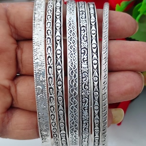 May include: A set of six silver-toned metal bangles featuring various engraved patterns, including geometric shapes, intricate floral-like motifs, and linear textures. Each bracelet has a distinct design with oxidised detailing to highlight the engravings.