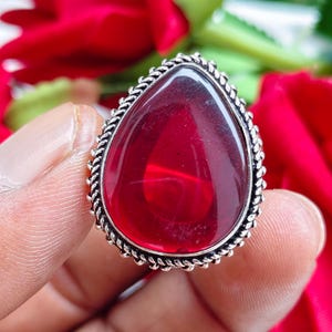Könnte beinhalten: Ein tropfenförmiger Ring mit einem großen, glatten, durchscheinenden tiefroten Stein, der in ein poliertes silberfarbenes Metallband gefasst ist. Die Fassung ist am äußeren Rand mit einer gedrehten, seilartigen Textur detailliert gestaltet.