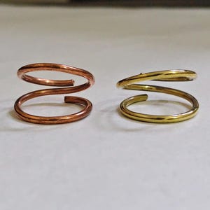 Könnte beinhalten: Zwei Spiralringe aus Draht, einer mit einer rotbraunen Kupferoberfläche und der andere mit einer glänzenden goldfarbenen Oberfläche. Jeder Ring weist ein durchgehendes gewickeltes Design mit zwei kreisförmigen Schlaufen auf, die sich leicht überlappen.