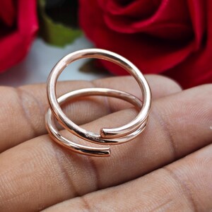 May include: A rose gold-toned bypass ring featuring a smooth, polished finish. The design consists of two overlapping bands that wrap around the finger, creating an open, adjustable silhouette with rounded, blunt ends and a reflective metallic surface.