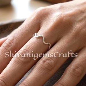 Puede incluir: Un anillo plateado con un diseño ondulado se lleva en un dedo. El anillo es el punto focal, sobre una superficie de madera. El texto "ShivanigemsCrafts" es visible en la superficie.