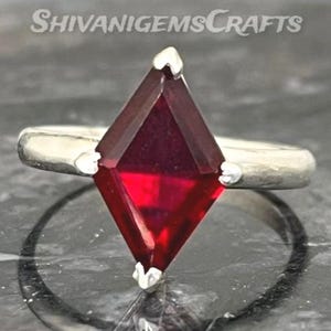 Könnte beinhalten: Ein silberner Ring mit einem drachenförmigen, tiefroten Edelstein, der von vier Krappen gehalten wird. Die Ringschiene ist glatt und metallisch. Der Text SHIVANIGEMSCRAFTS ist oben sichtbar. Das Design ist zentriert und symmetrisch.