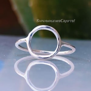 Könnte beinhalten: Ein silberner Ring mit kreisförmigem Design. Der Ring hat einen großen offenen Kreis als Hauptelement, mit einem dünnen Band, das sich zu den Seiten erstreckt. Der Ring wird auf einer reflektierenden Oberfläche präsentiert. Der Text "ShivanigemsCrafts!" ist im Hintergrund sichtbar.
