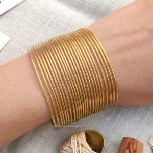 Könnte beinhalten: Ein Set aus einundzwanzig dünnen, goldfarbenen Metallarmreifen, die zusammen als ein breites Manschettenarmband getragen werden. Die Armreifen haben eine glatte, polierte Oberfläche und eine kreisförmige Form, was einen Stapel-Look am Handgelenk erzeugt.