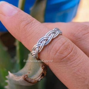 Könnte beinhalten: Ein silberfarbener Ring mit einem geflochtenen Drahtdesign, das sich um das Band legt. Das Metall hat eine polierte Oberfläche und ein durchgehendes, gewebtes Muster. Der Text ShivaniGemsCrafts ist auf der Oberfläche des Ringbandes sichtbar.