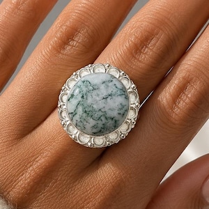 Könnte beinhalten: Ein Silberring mit einem großen, runden Moosachat-Cabochon mit grünen dendritischen Einschlüssen. Der Stein ist in eine dekorative, gewellte Silberfassung eingefasst, die am Rand mit einer Reihe kleiner, erhabener, runder Perlen verziert ist.
