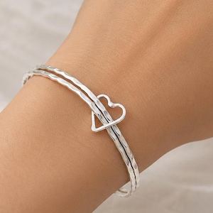 May include: A silver-toned bangle bracelet featuring a textured, hammered surface and a central open heart-shaped charm. The bracelet consists of two thin, parallel bands that meet at the heart detail, creating a delicate and continuous circular design.
