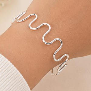Könnte beinhalten: Ein silberfarbenes Armband mit einem durchgehenden, fließenden, wellenförmigen Liniendesign, das sich um das Handgelenk legt. Das Metall hat eine glatte, polierte Oberfläche und bildet ein einfaches, sich wiederholendes Schlangenmuster.