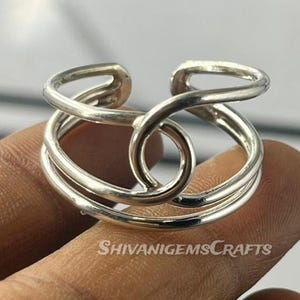 Könnte beinhalten: Ein silberfarbener Ring mit einem einzigartigen offenen Design. Der Ring hat eine Reihe von geschwungenen Linien und ein zentrales kreisförmiges Element. Der Markenname "ShivanigemsCrafts" ist sichtbar.