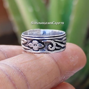 Könnte beinhalten: Ein silberner Ring mit floralem und Ranken-Design. Der Ring hat ein zentrales Blumenmotiv, das von detaillierten Ranken umgeben ist. Der Ring wird im Nahaufnahme gezeigt, um die Details hervorzuheben. Der Ring hat die Größe 54.