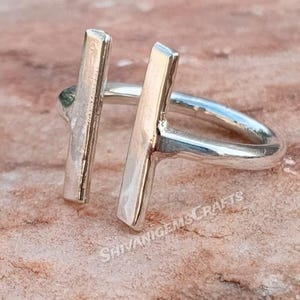 Könnte beinhalten: Nahaufnahme eines silbernen Rings mit einem einzigartigen T-Bar-Design. Der Ring besteht aus poliertem Metall und verfügt über zwei vertikale Stäbe, die durch ein horizontales Band verbunden sind. Der Ring befindet sich auf einer strukturierten, braunen Oberfläche. Der Text "SHIVANIGEMSCRAFTS" ist sichtbar.