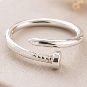 Könnte beinhalten: Ein polierter, silberfarbener Ring in Form eines gebogenen Nagels. Das Band hat eine glatte, abgerundete Oberfläche, die sich um den Finger legt, mit einem ausgeprägten Nagelkopf und strukturierten, gerillten Details am Ende des Metalls.