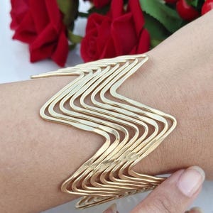 Könnte beinhalten: Ein goldfarbenes Armband in Manschettenform, bestehend aus einer Reihe paralleler, zickzackförmiger Metalldrähte, die ein geschichtetes, wellenartiges Muster bilden. Die Metallstruktur ist offen und flexibel und umschließt das Handgelenk.