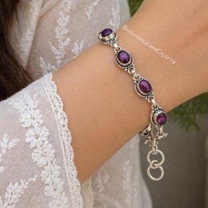 Könnte beinhalten: Ein silbernes Kettenarmband mit vier ovalen, tiefvioletten Cabochon-Edelsteinen in verzierten Metallfassungen. Das Armband verfügt über einen kleinen runden Verschluss und einen hängenden Ring, wobei der Text SHIVANIGEMSCRAFTS auf der Haut sichtbar ist.