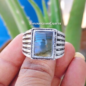 Könnte beinhalten: Ein silberner Ring mit einem rechteckigen Labradoritstein. Der Stein hat blau-grüne schillernde Farben. Der Ring hat ein strukturiertes Band mit horizontalen Linien. Der Text "SHIVANIGEMSCRAFTS" ist im Hintergrund sichtbar.
