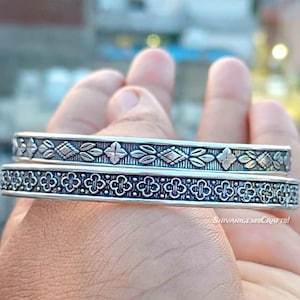 Könnte beinhalten: Zwei silberne Armreifen mit aufwendigen floralen und geometrischen Mustern. Der obere Armreif hat ein Design aus Blättern, Rauten und Kreuzen, während der untere ein sich wiederholendes kleeblattartiges Motiv aufweist. Die Armreifen werden in einer Hand gehalten.